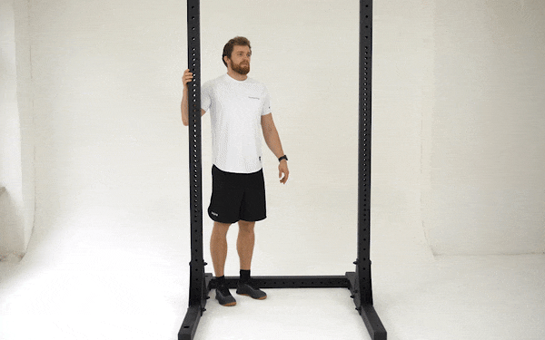 A GIF of a white man in a white t-shirt and black shorts stretching his chest. He stands inside a black metal exercise frame. He grips the frame with his right hand and takes two steps forward, letting his right arm stretch out behind himself. His elbow stays straight and his shoulder engaged to counteract the pull of the frame.