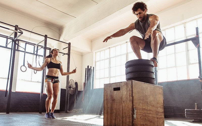 A white woman jumps rope while a man with curly brown hair does a box jump in a gym. They appear to be doing a couple workout of different activities.