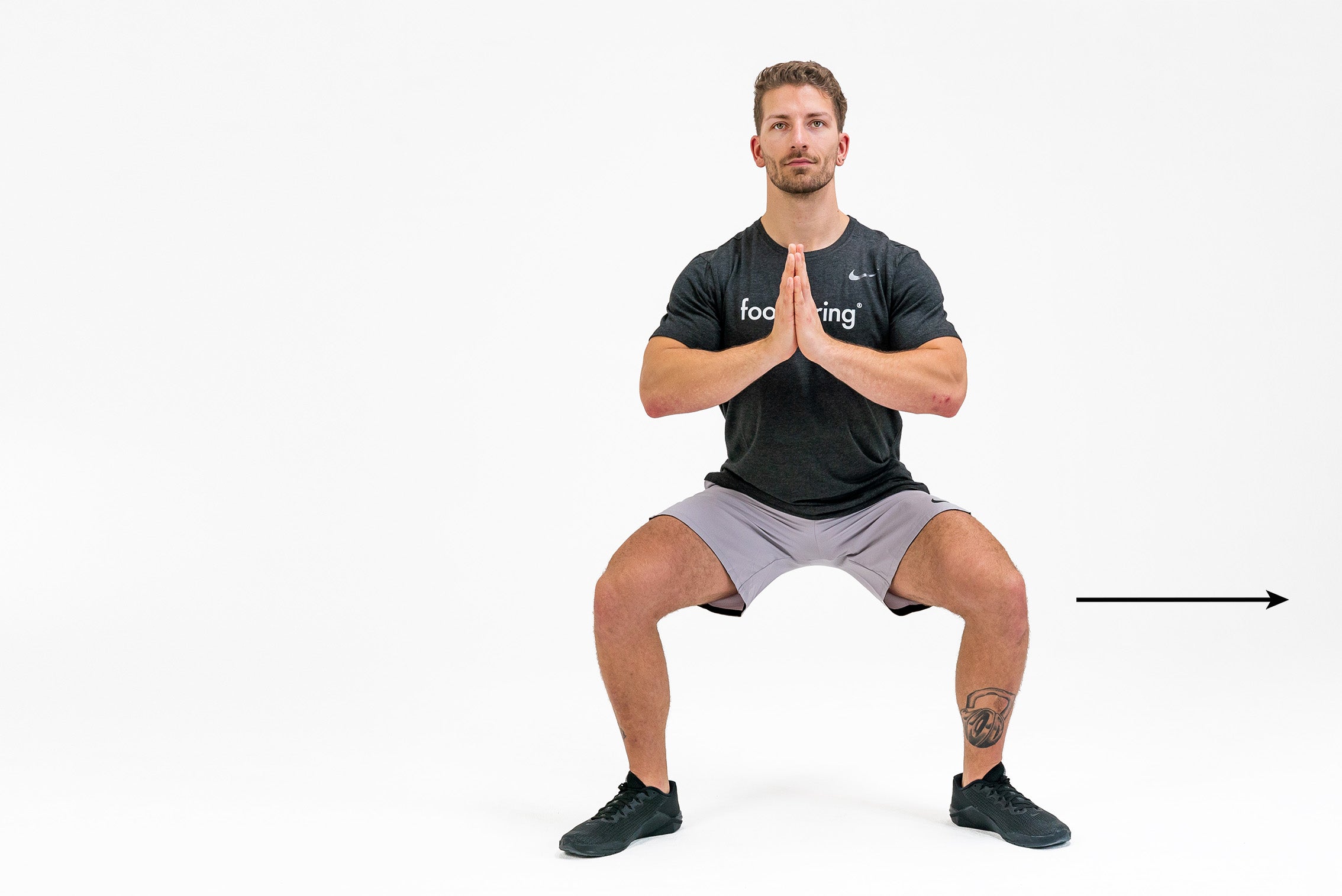 A white man in a black foodspring t-shirt demonstrates the position for a crab squat walk, while a black arrow points to his left indicating which direction he is about to go.