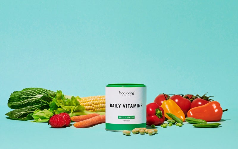 Daily Vitamins by foodspring sit with a selection of vegetables in front of a turquoise background