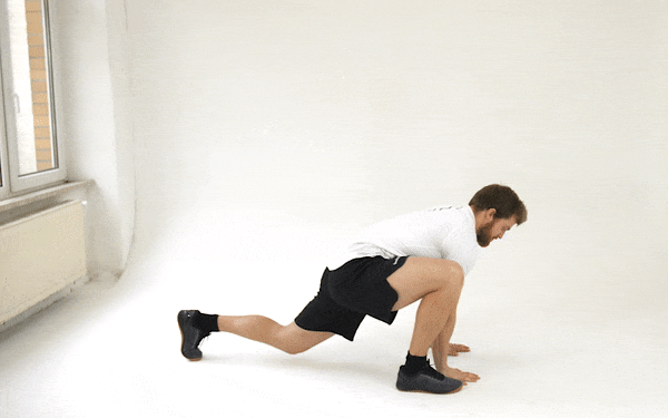 A GIF of a white man in a white t-shirt and black shorts doing a deep lunge with a shoulder rotation. He lunges his right leg forward until it is even with his left hand, which rests on the floor directly under his shoulder, while his left leg remains extended behind him, resting on the toes. He stretches his right hand towards the ceiling and looks in the same direction as the hand. Then he moves his right hand to the floor, between his right foot and left hand. He brings his right foot behind him to a plank position, then brings his left foot forward into a lunge next to his hands. He stretches his left arm straight upwards, and follows the movement with his gaze again. 