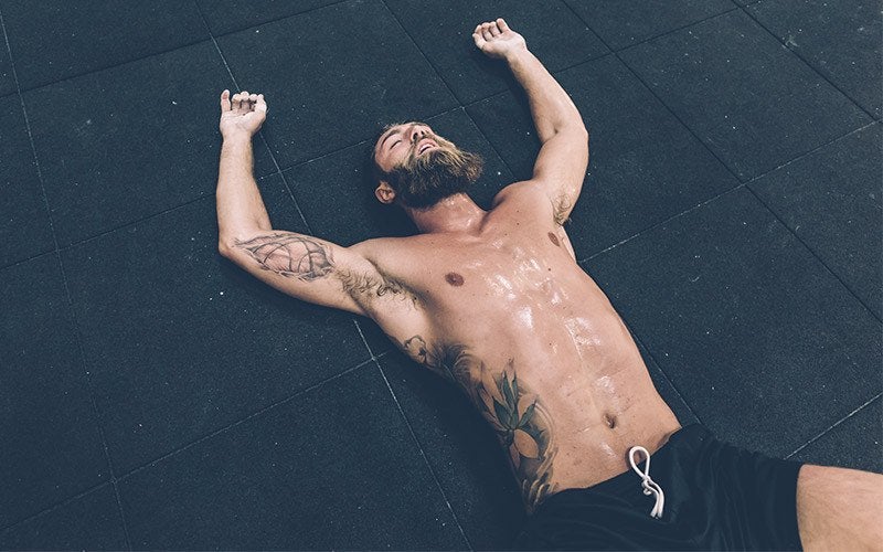 A shirtless, tattooed, bearded white man appears to be lying on the floor in exhaustion, with his torso covered in sweat.