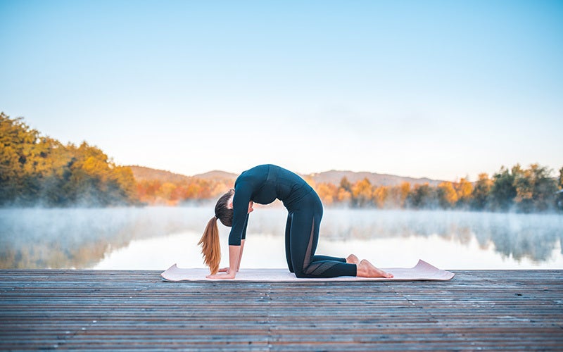 A white woman on a yoga mat on a pier at a tree-lined lake with mist rising up from the water performs a cat-cow stretch