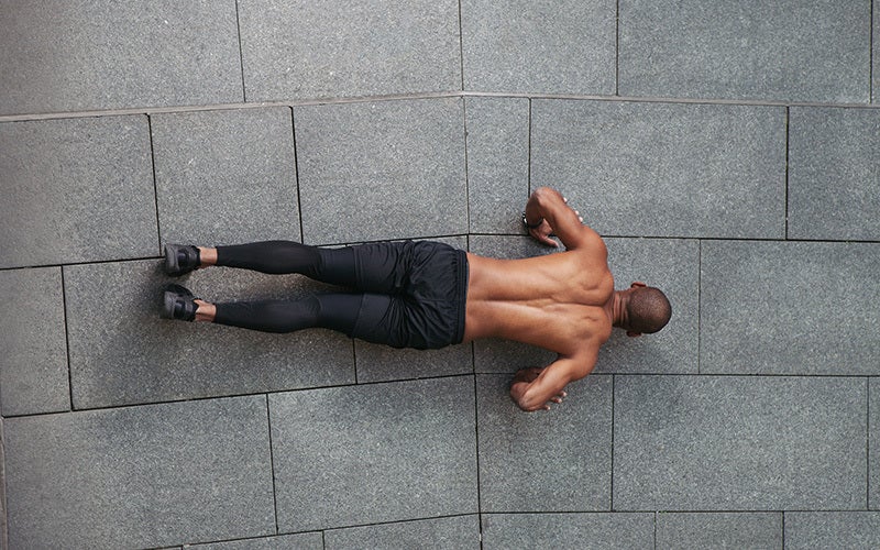 A man of color with black pants and no shirt is seen from above performing a push-up on gray granite tiles outdoors
