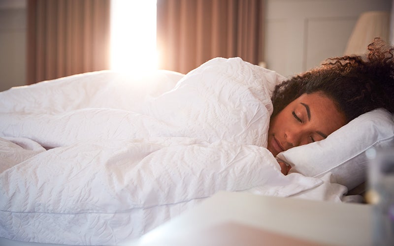 A woman of color sleeps on a white pillow and is snuggled under a white duvet. Sunlight washes in from the partly opened tan curtains behind her.