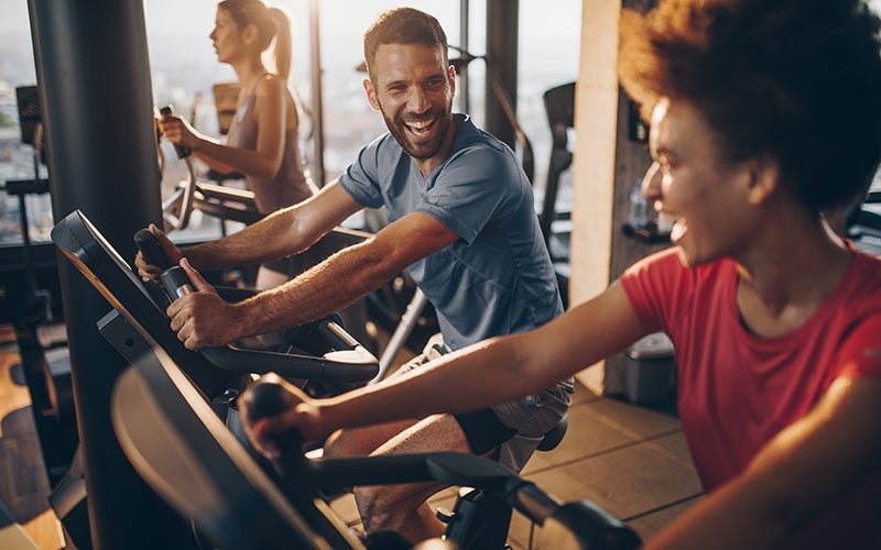 a white man and a woman of color smile at each other while riding stationary bikes in a gym setting. In the background, a white woman appears to be using an elliptical machine.