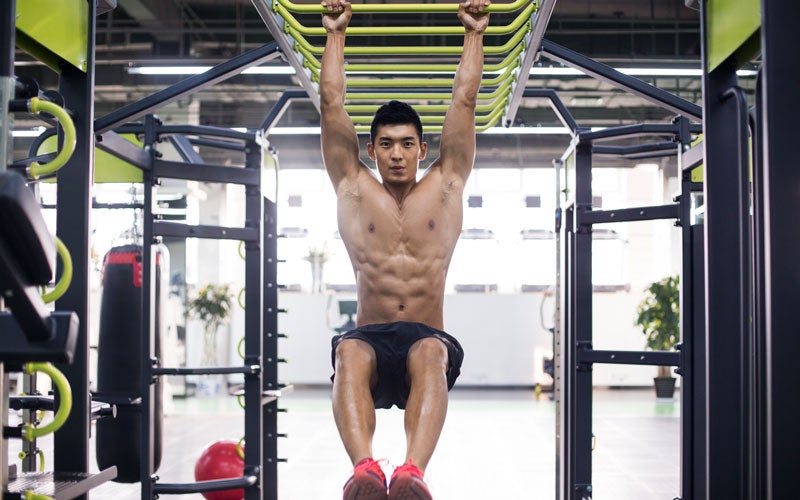 A shirtless man with Asian features hangs off of a set of monkey bars in a gym, staring at the camera.
