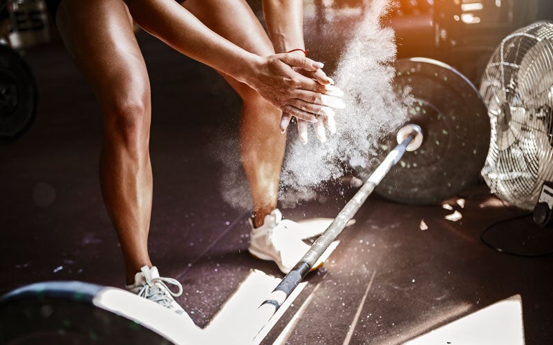A pair of arms and shaved legs in front of a barbell. The hands are clapping away excess chalk dust before a grip strength exercise.