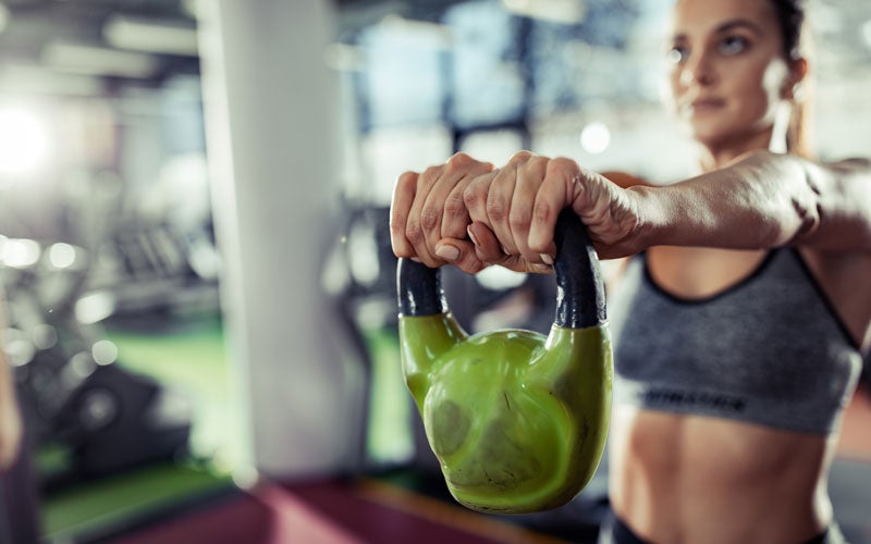 A white woman is out of focus in the photo, holding a green kettlebell in tight focus in the foreground.