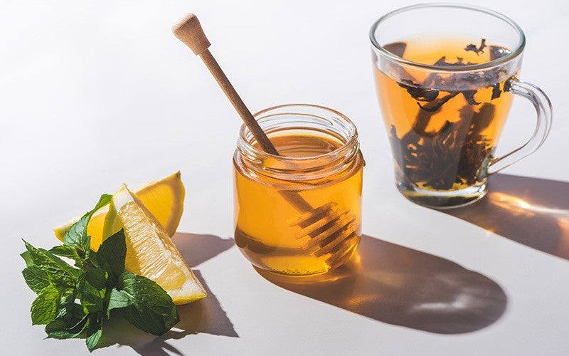 Photo of a glass mug with loose tea leaves floating in it, a jar of honey with a honey dipper, two lemon slices and a sprig of mint