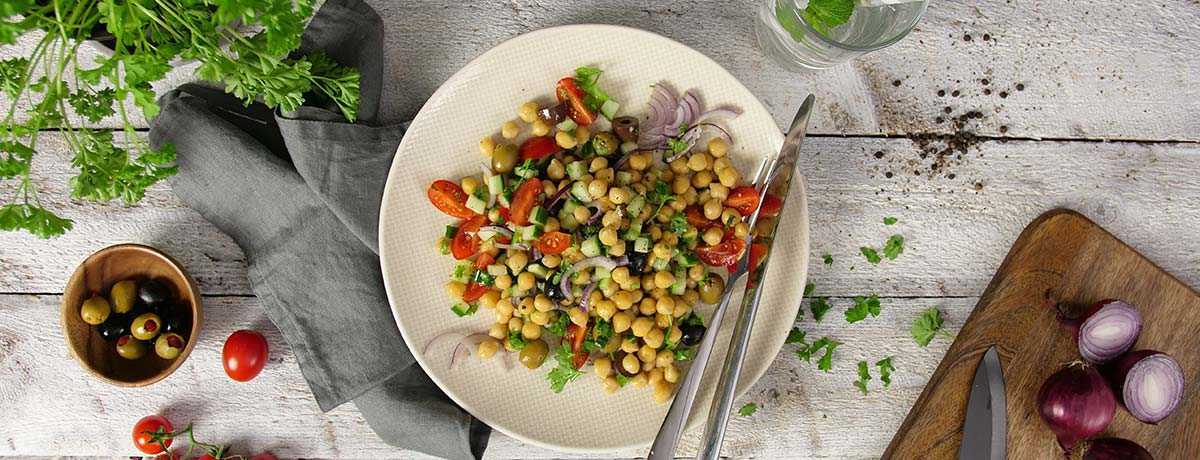 A white plate holds a chickpea salad with bright accents of tomatoes, red onions, cucumber cubes, and coriander leaves