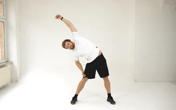 A GIF of a white man in a white t-shirt and black shorts performing lateral stretches. He stands with his feet wide apart. He leans first to his left, resting his left hand on his thigh and raising his right hand above, then past, his head. He then straightens up and does the same on his right side.
