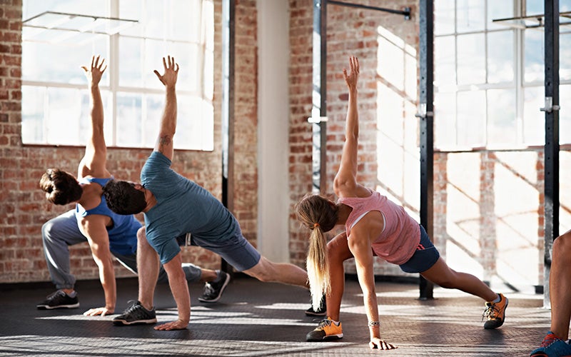 Three athletes in sports gear inside a brick-walled gym do a deep lunge/warrior stretch, with one hand on the floor and the other reaching up high to the ceiling.