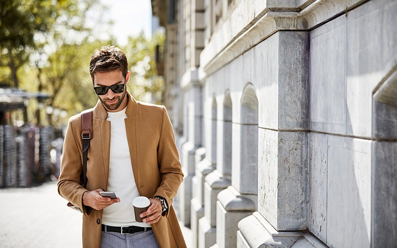 Mann läuft mit Kaffee in der Hand durch die Stadt und schaut dabei auf sein Handy