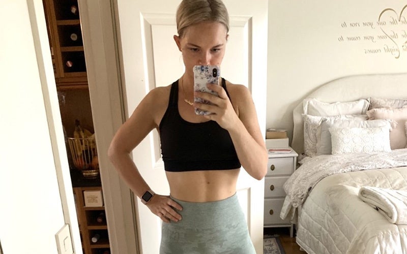 A selfie of Nathalie, a white woman in a black sports bra and gray high-waisted pants, showing her feel-good body with a defined line in the middle of her abs. She stands in front of a mirror, holding her phone in front of herself, and the background shows the edge of a bed and a white nightstand, as well as a white door.