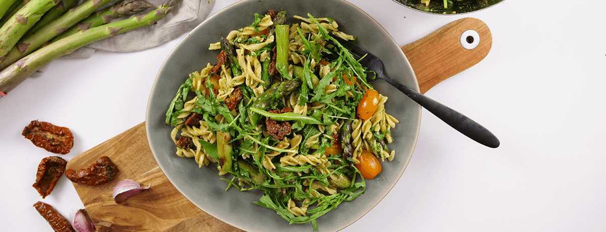 A gray bowl holds pasta salad with asparagus and pesto, garnished by dried and fresh tomatoes.