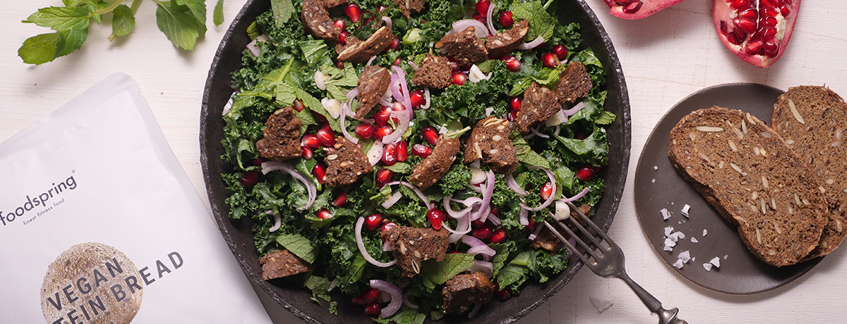 A vibrant green kale salad with bright red pomegranate seeds and brown Protein Bread croutons