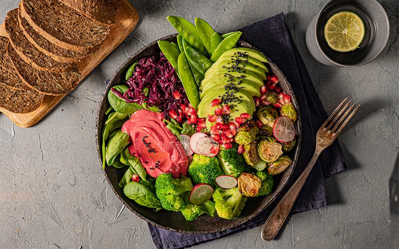 Photo from above of a Buddha bowl with broccoli, pink-tinted beetroot hummus, roasted Brussels sprouts, avocado, radish slices, purple cabbage, green snap peas and avocado slices, topped with pomegranate seeds and chia seeds. Next to the bowl is a stomeware mug of tea topped with a round slice of lime, and a wooden board spread with slices of Protein Bread.