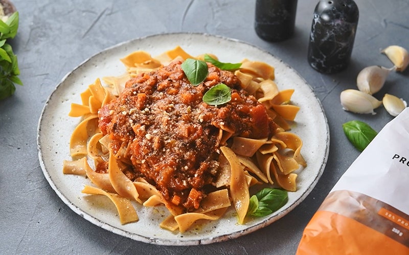 A plate of foodspring's Protein Pasta with a red bolognese sauce on top, garnished with basil leaves and sprinkles of Parmesan cheese