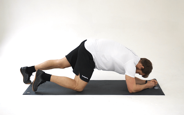 a GIF image of a white, short-brown-haired man in sports gear getting himself into the basic plank position. He supports himself on elbows and knees, then walks his legs out one by one to balance on his toes, and supports the weight of his upper body on his forearms, which are flat on his exercise mat with his elbows directly under his shoulders. His upper and lower body should form a straight line with his pelvis kept low and stable. 