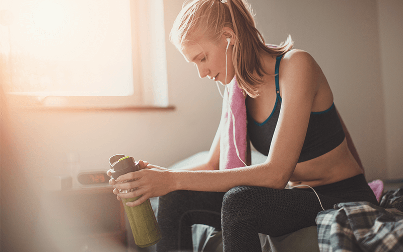 Sportliche junge Frau sitzt nach dem Training mit Sportkleidung und grünem Smoothie auf dem Bett
