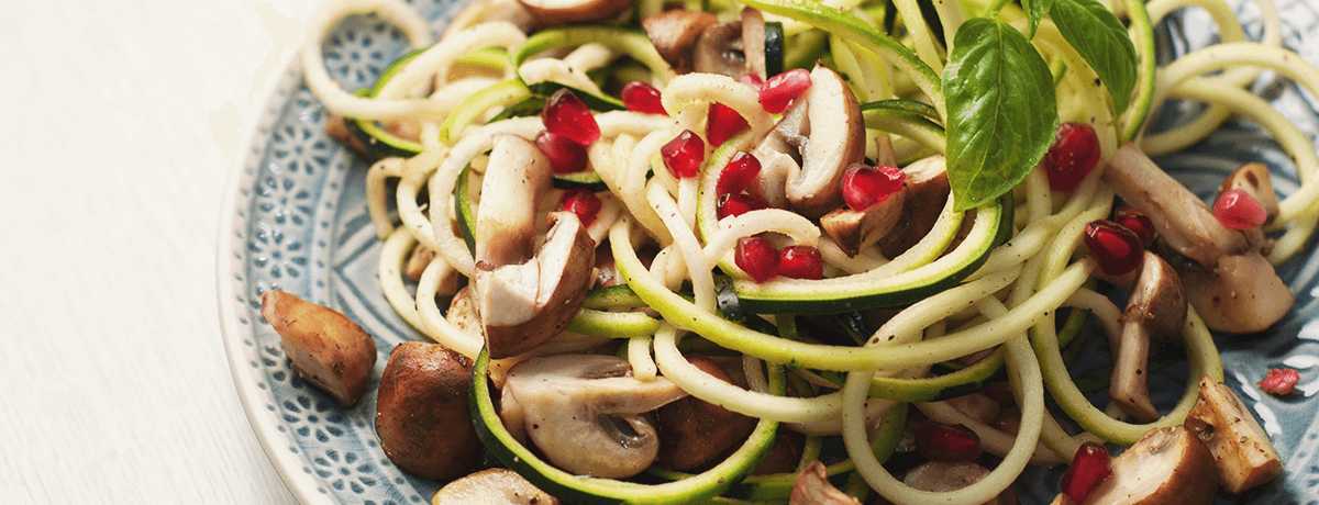 A photo of courgetti/zoodles with mushrooms and pomegranate seeds on a plate with a blue and white pattern and garnished with green herbs