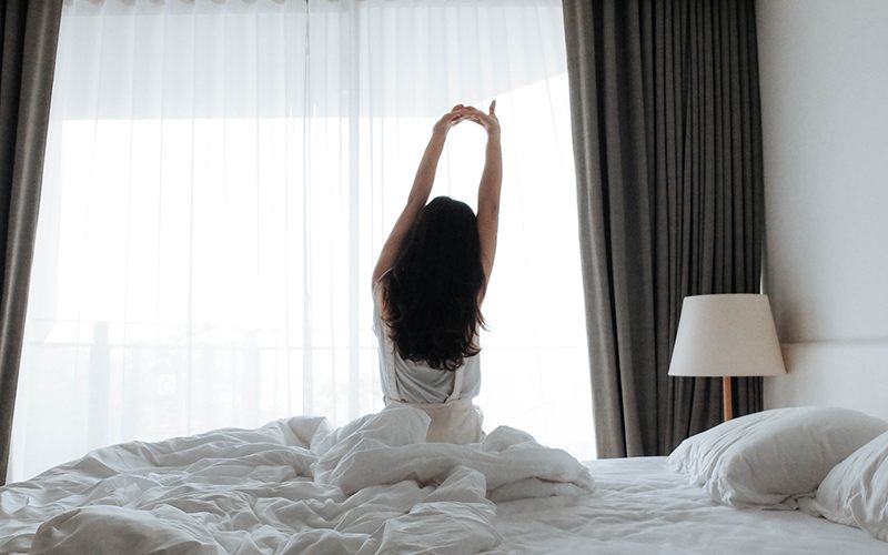 A long-haired person seen from behind does morning stretches in their bed, stretching their arms above their head.