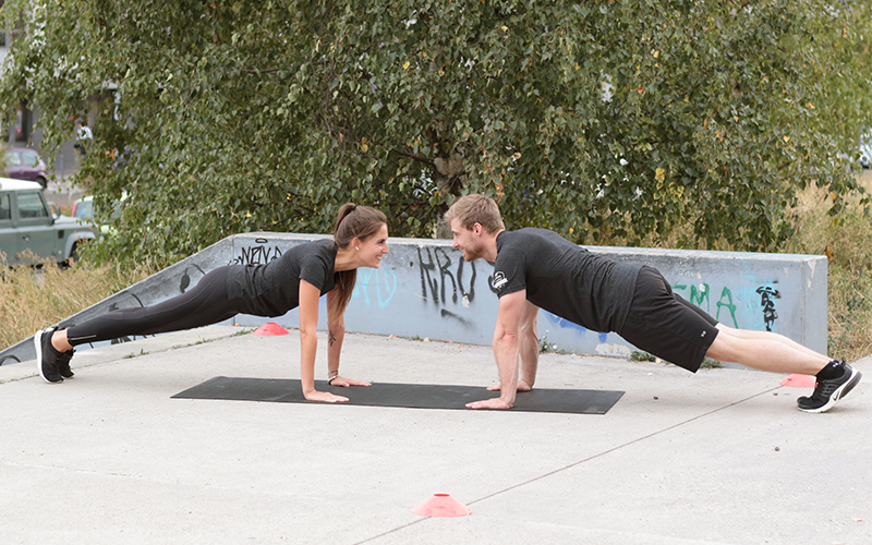 A white man and a white woman doing a partner workout of planks outdoors