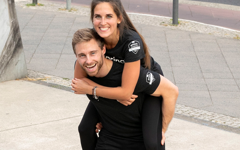 A white woman rides piggyback on a white man's backpack on a sidewalk