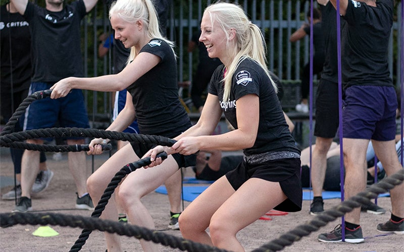 Two blonde-haired white women doing a partner workout with large black ropes