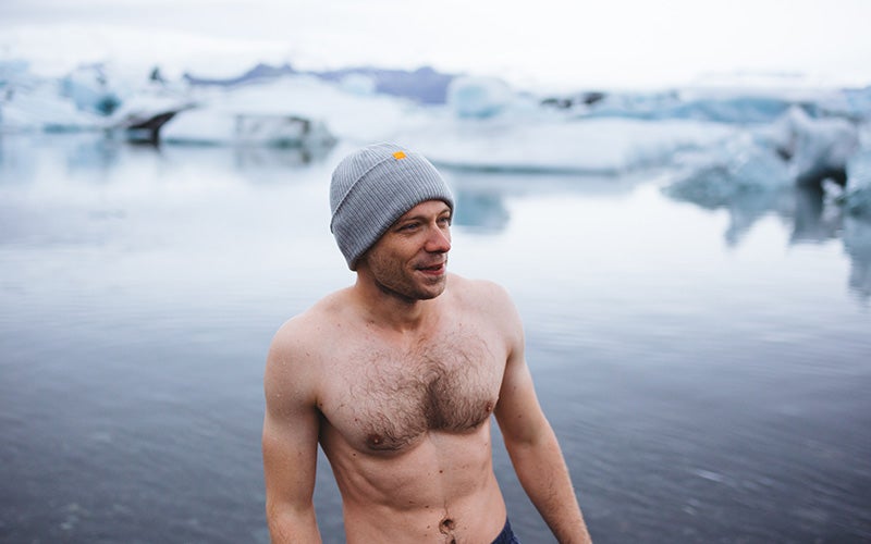 Uomo in piedi a petto nudo e con un berretto di lana in testa davanti a un lago quasi ghiacciato