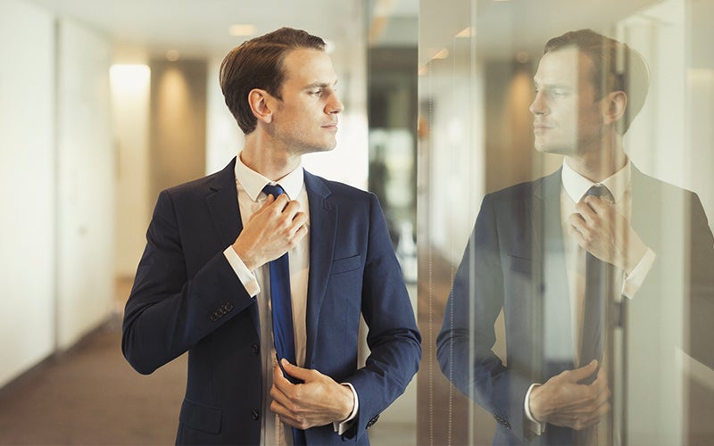 A white man in a suit adjusts his tie, while checking his reflection in a window which is acting as a mirror