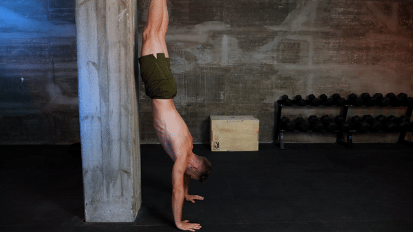 A white shirtless male athlete does a handstand, alternating lifting one hand off the floor to touch the opposite shoulder, then switching sides.