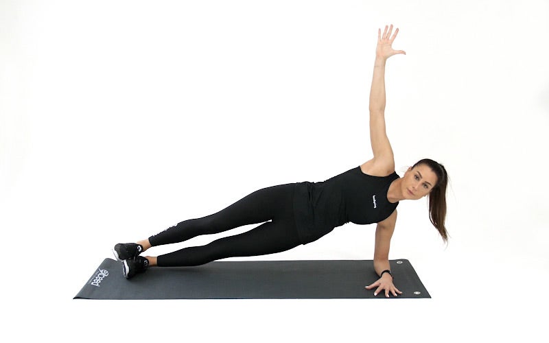 A light-skinned woman in black workout gear with a brown ponytail demonstrates a side plank: balancing on her left foot and left forearm, she raises her right arm into the air. Her body is at a diagonal downward angle.