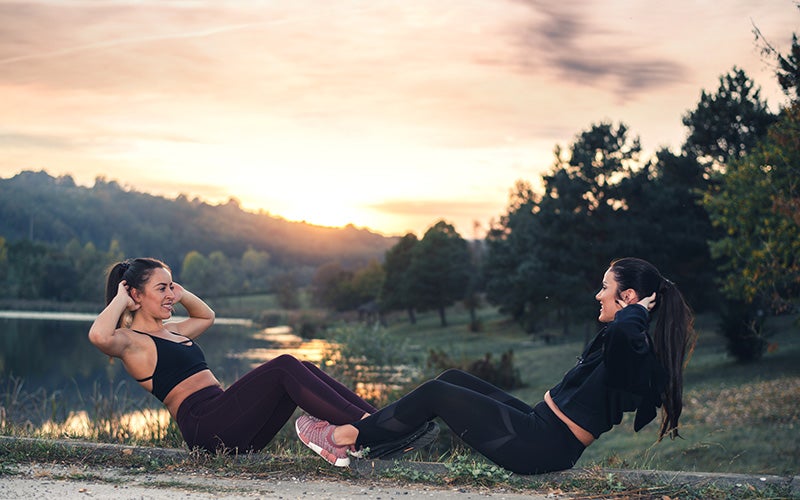 two white-presenting women with dark brown ponytails do sit-ups in front of a lake with the sun setting behind them