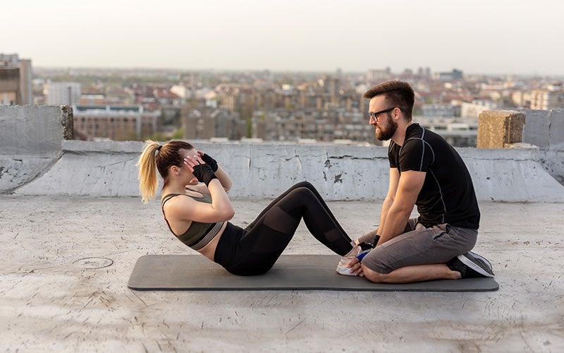 A white woman does sit-ups on a mat outdoors on what appears to be a roof terrace in a city background. A man in glasses holds her feet down.