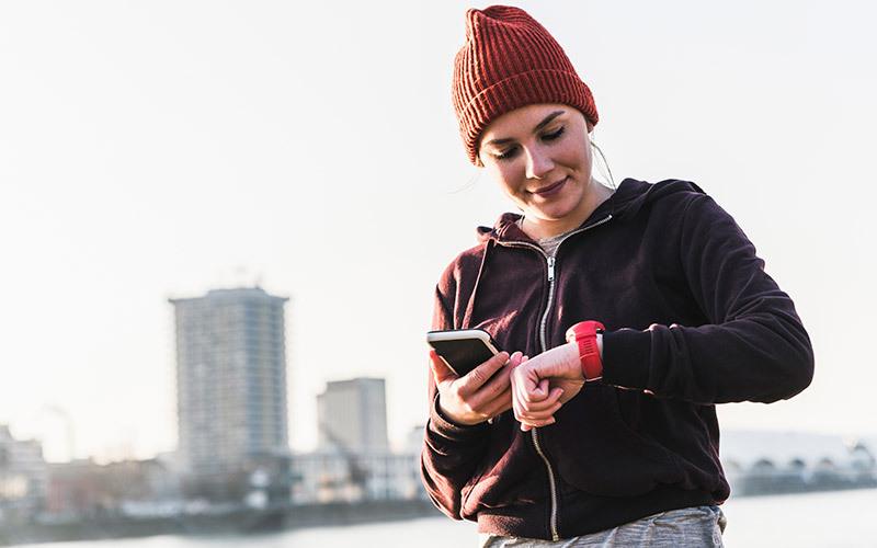A white woman in a knit red beanie with a city background behind her checks her bright red sports watch, holding her phone in her other hand to keep a workout log.