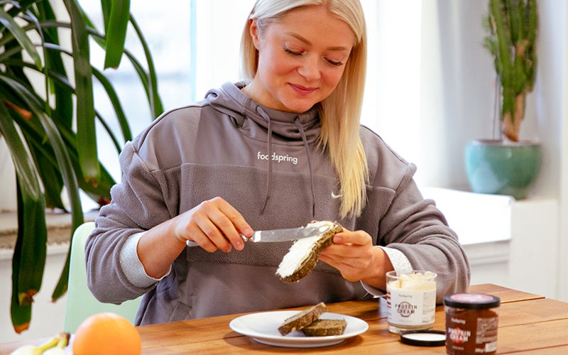A white, blonde-haired woman in a gray foodspring-branded sweatshirt practices IIFYM while spreading coconut-flavored Protein Cream on a slice of bread.