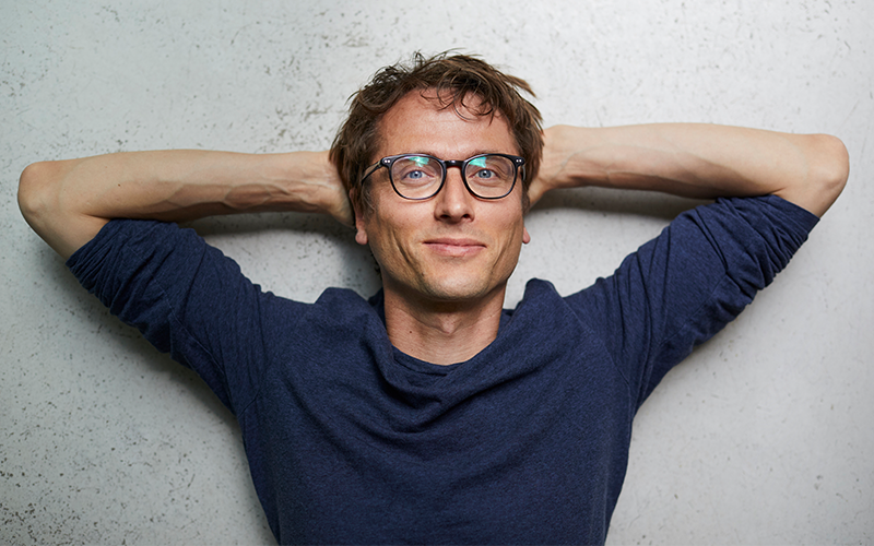 A short-haired masculine-presenting person wearing black-framed glasses stands in front of a wall with a slight smile, meditating on contentment