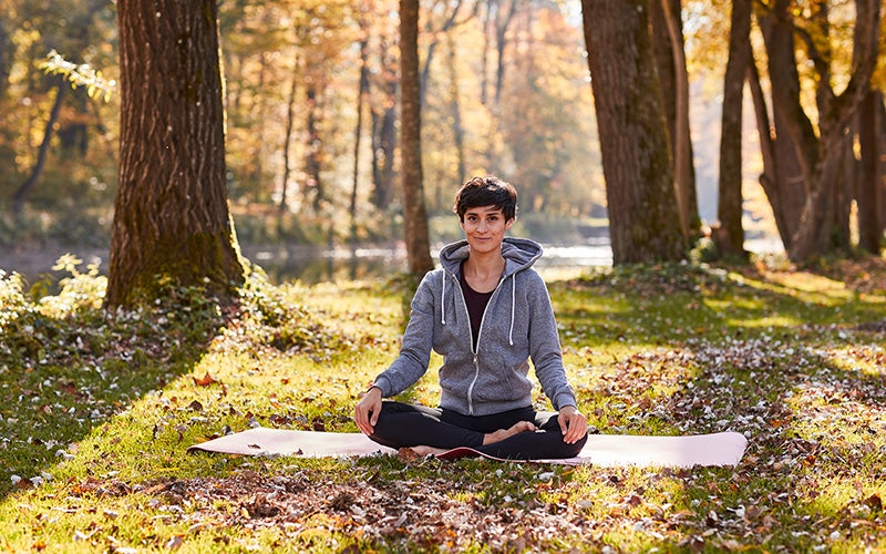 A short-haired white woman sits on a yoga mat in the forest.