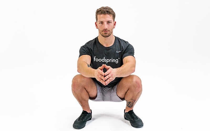 a white man in a black foodspring t-shirt rests in a squat position, looking straight into the camera