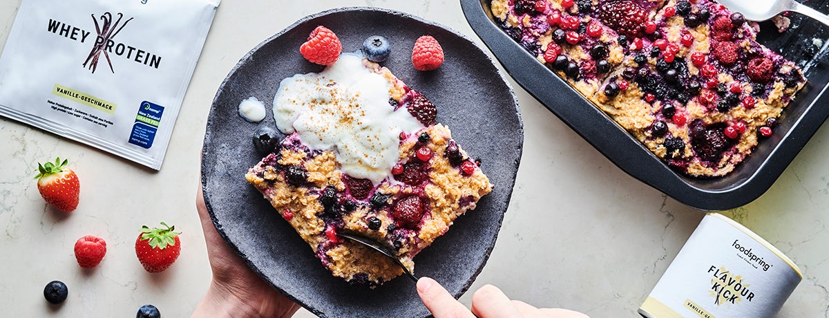 A hand digs a spoon into a portion of berry-loaded baked oatmeal, with a packet of Whey Protein on the side. 
