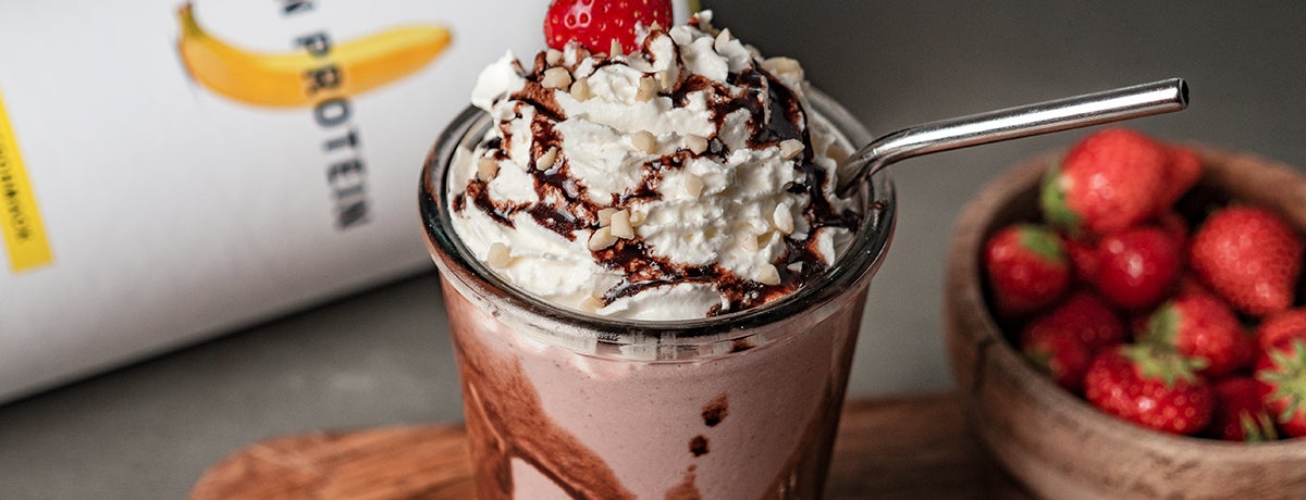 A glass drizzled with chocolate sauce, filled with pink smoothie, topped with a mountain of whipped cream, nuts and chocolate sauce, all with a strawberry on its peak. There is a bowl of fresh strawberries to one side and an out-of-focus canister of banana-flavored Vegan Protein on the other, making the most of pea protein.
