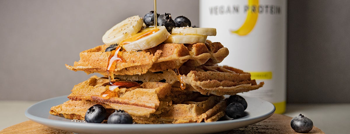 A stack of golden-brown, fluffy vegan banana waffles garnished with banana slices, blueberries, and maple syrup, with a canister of banana Vegan Protein behind it