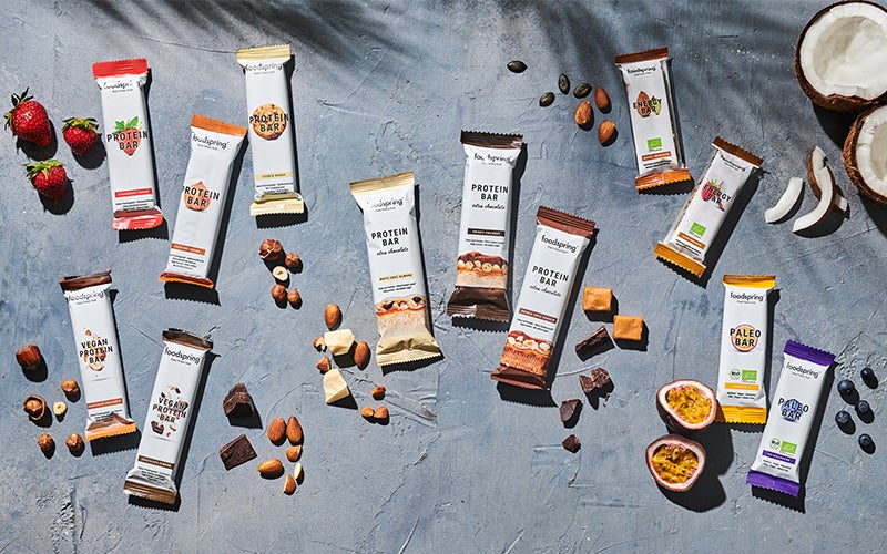 A selection of foodspring's best energy and protein bars laid out on a gray background with ingredients such as nuts, chocolate chunks, and caramel strewn artistically around them.