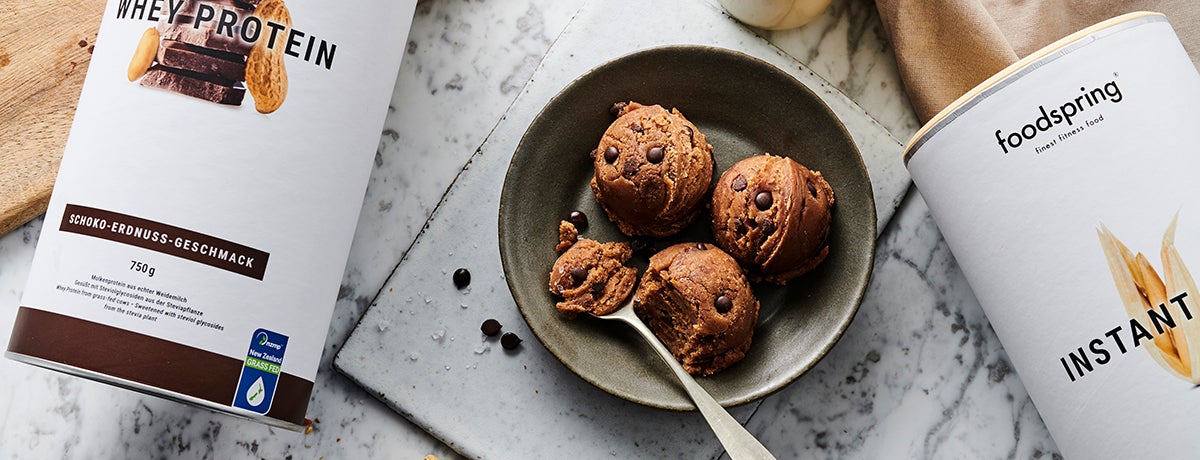 A spoon scoops into a bowl of chocolate chip cookie dough next to a canister of chocolate peanut butter Whey Protein and another of Instant Oats by foodspring.