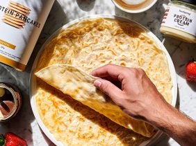 A medium-skin-tone hand stretches over a batch of Protein Crepes with a background of fresh red berries, a glass of water, and a canister of foodspring's Protein Pancakes.