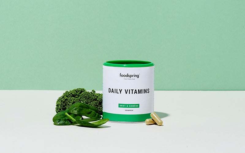 a canister of foodspring's Daily Vitamins in front of a green background, sitting next to a bright green head of broccoli.