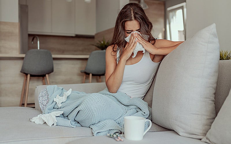 Frau sitzt mit Erkältung mit Bett und putzt sich die Nase 