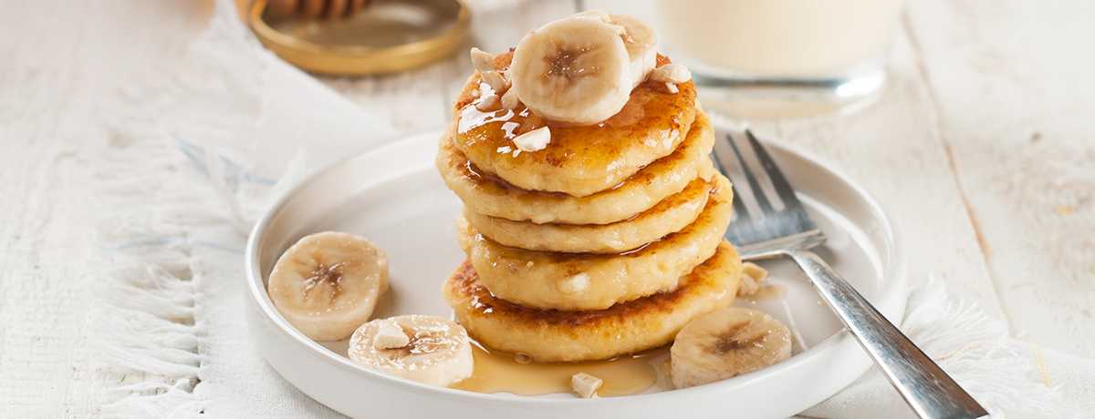 A stack of Protein Pancakes topped with syrup and banana slices sits on a white plate with a silver fork lying next to it